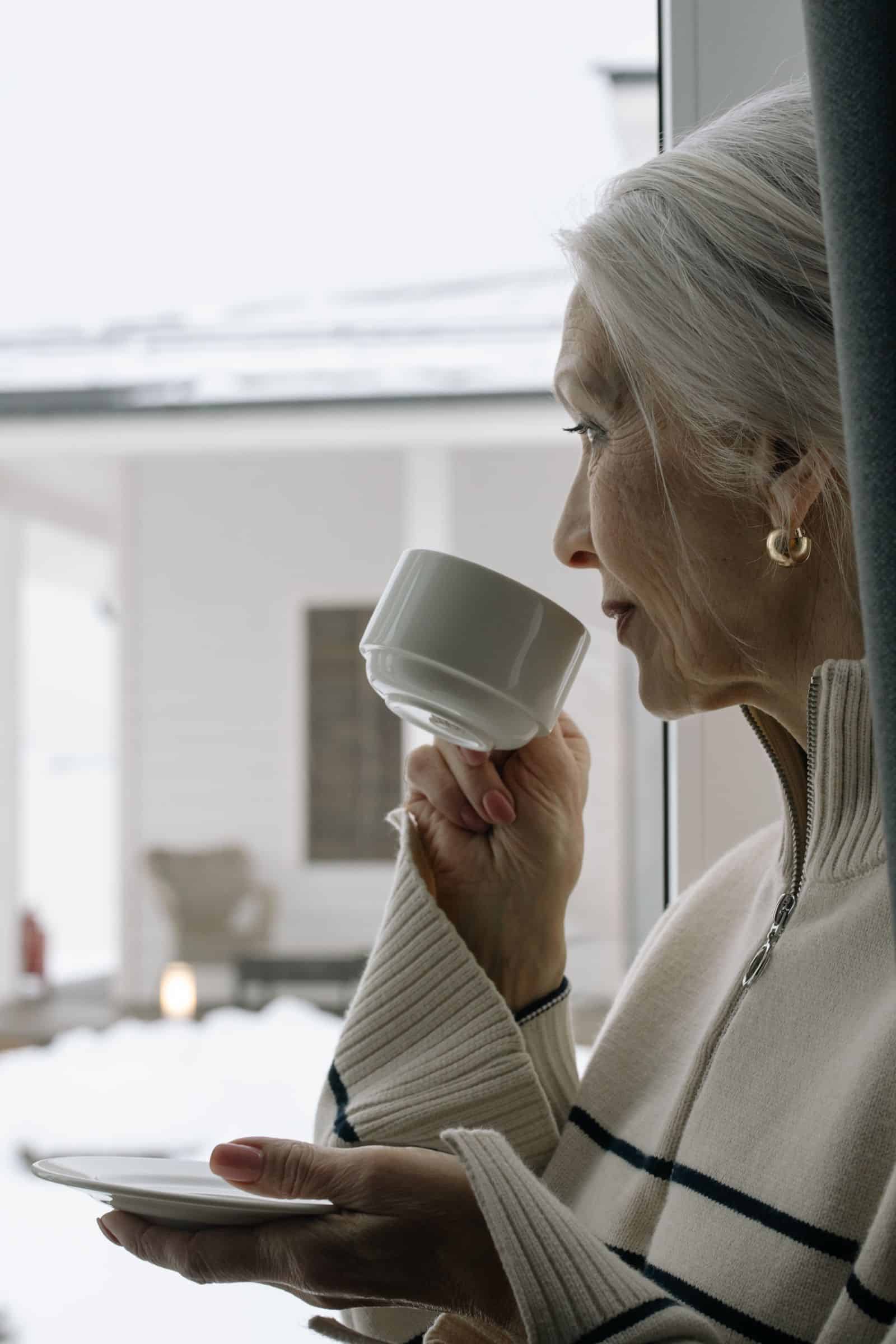 Older woman with a warm drink looking out at a snowy window